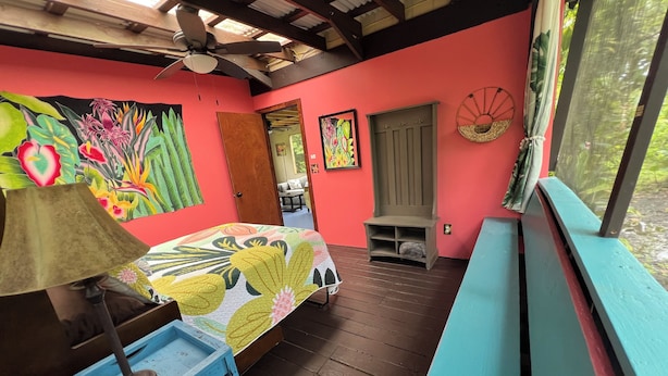 Una habitación con cama, ventilador de techo, banco de madera y un mural colorido en la pared.