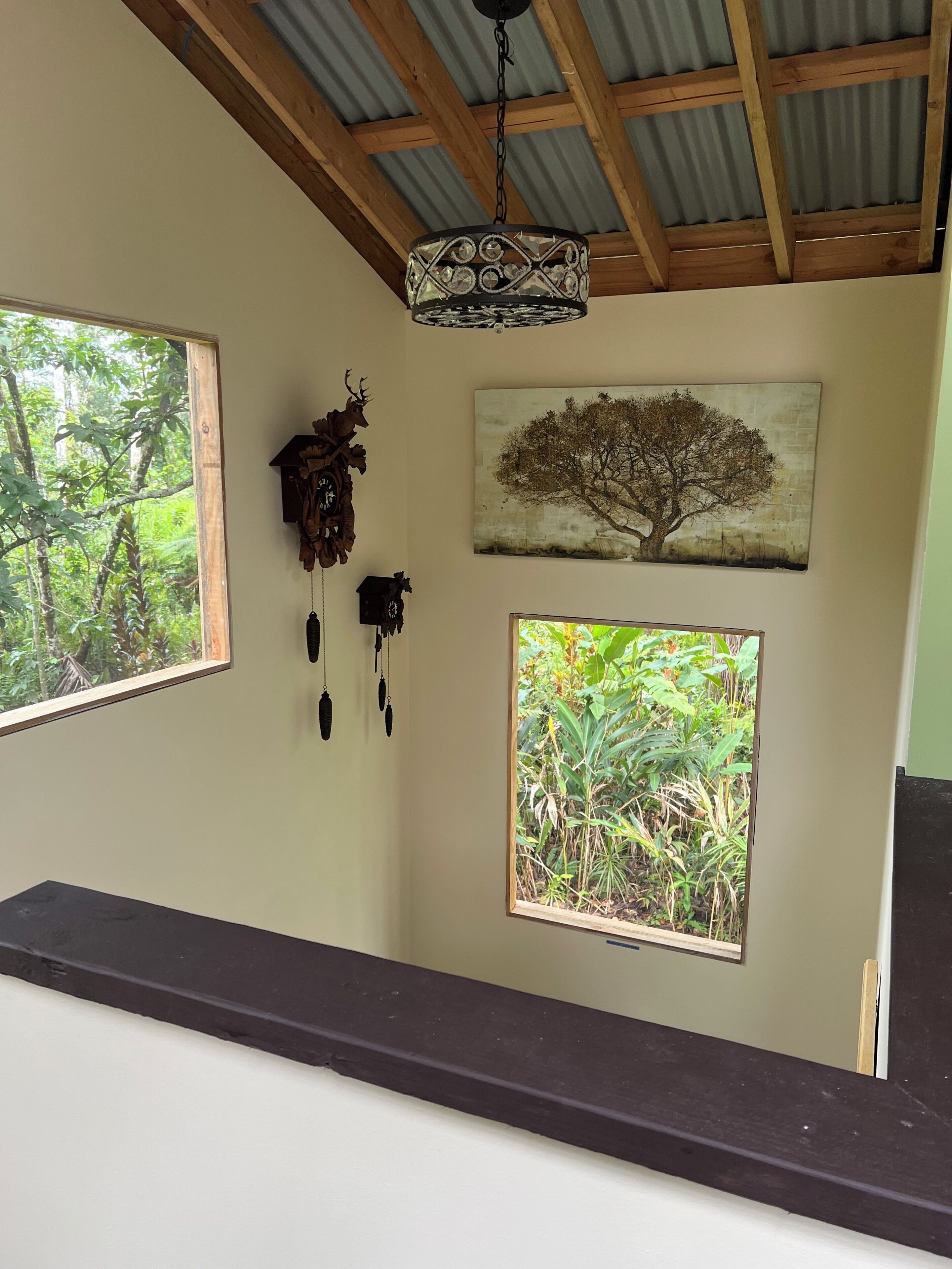 Ein Zimmer mit Holzboden, einer Pendelleuchte, einer an der Wand befestigten Kuckucksuhr, einem Bilderrahmen mit Baumdarstellung und einem Fenster mit Blick ins Grüne.