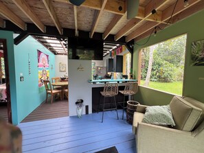 A room with a wooden ceiling, a dining area with chairs and a table, a kitchen with a counter and chairs, and a window with a view of greenery.