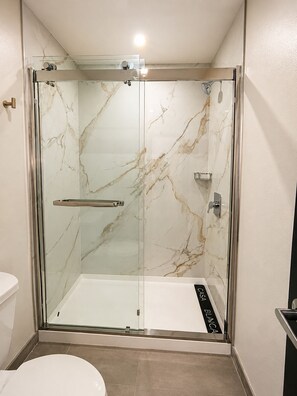 Aseo con ducha envuelta en cristal, pared de mármol, puerta de cristal y panel de control.