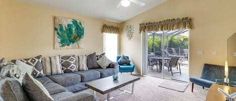 A living room with a sofa, coffee table, and a blue armchair. There is a ceiling fan, a painting, and a view through a glass door to an outdoor patio.