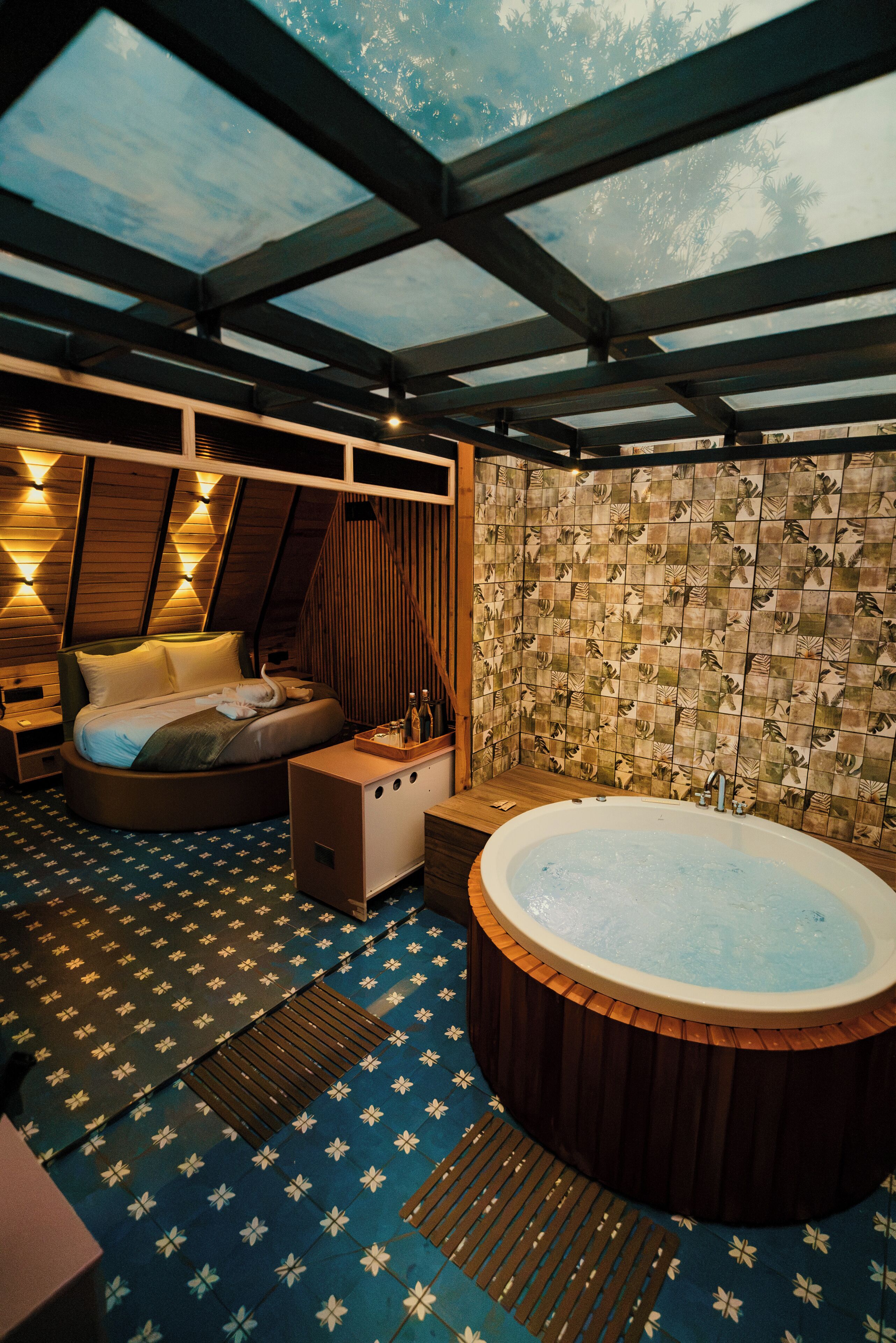 A room with a bed, a wooden bench, a hot tub, and a wall with patterned tiles.