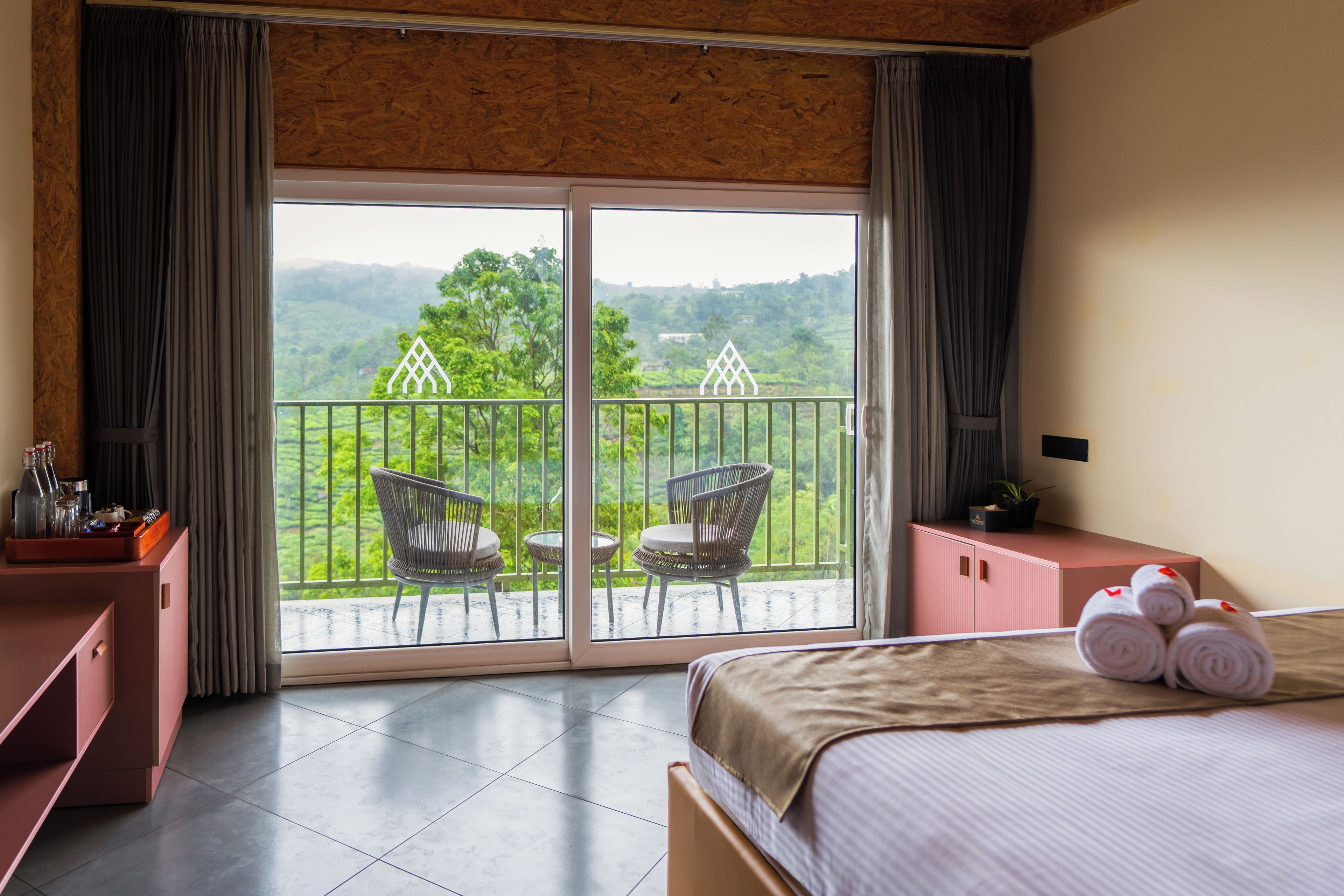 A hotel room with a bed, a small table with a bottle and glasses, a chair, a cabinet, and a balcony with a view of greenery.