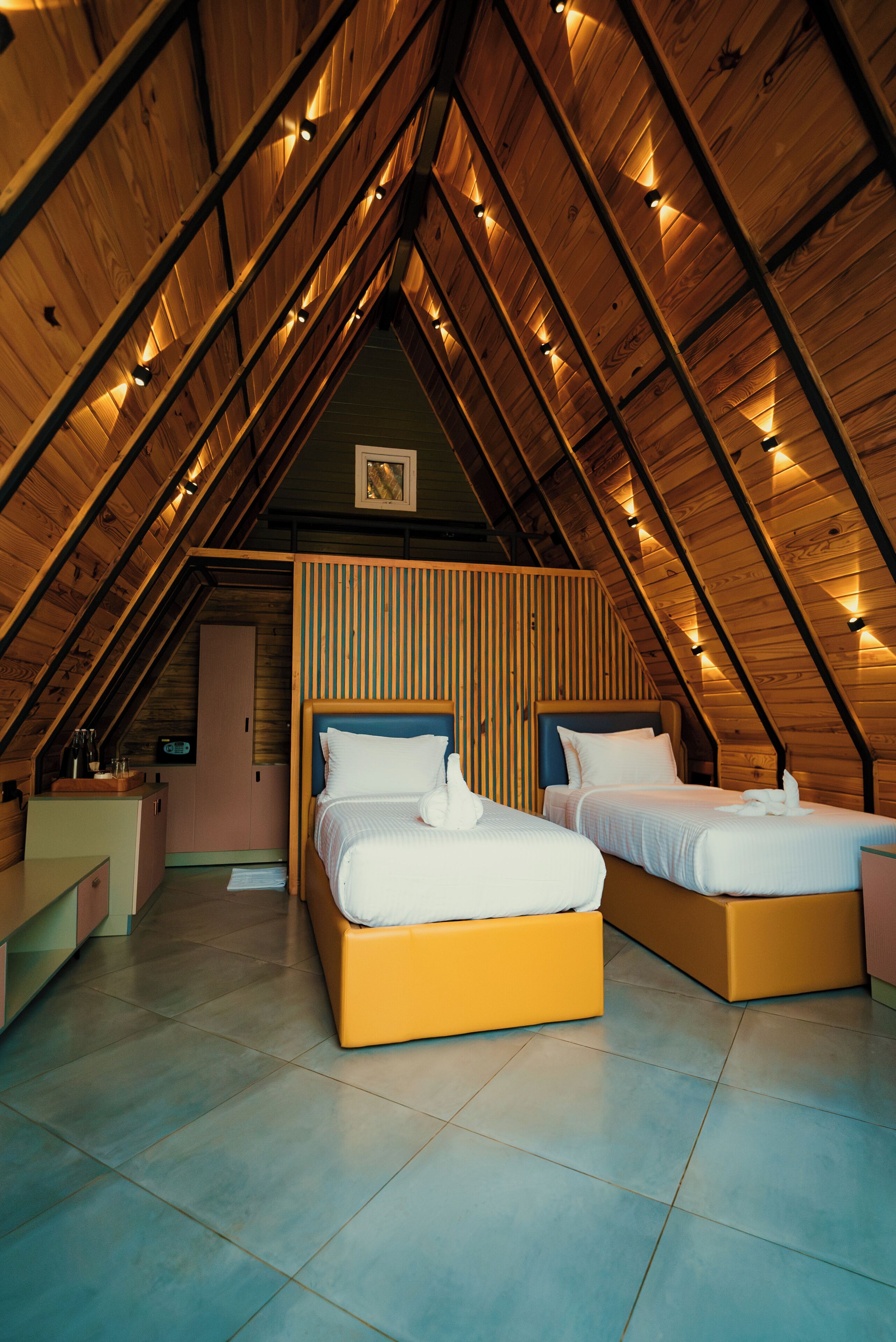 A cozy attic bedroom with two beds, a kitchenette, and a sloped ceiling with wooden beams.