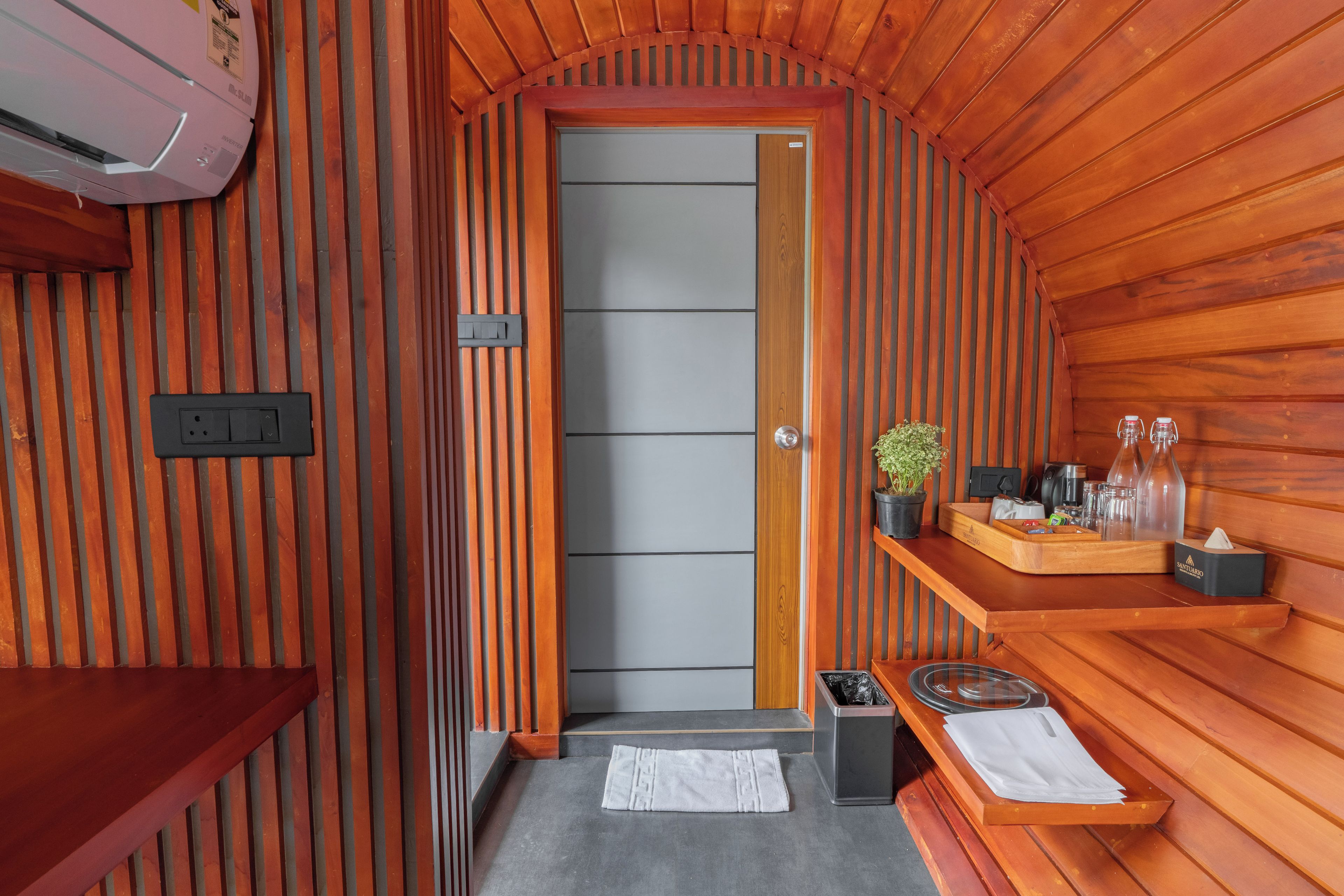 A compact, wooden interior with a door, a small trash bin, a towel, and a shelf with toiletries.