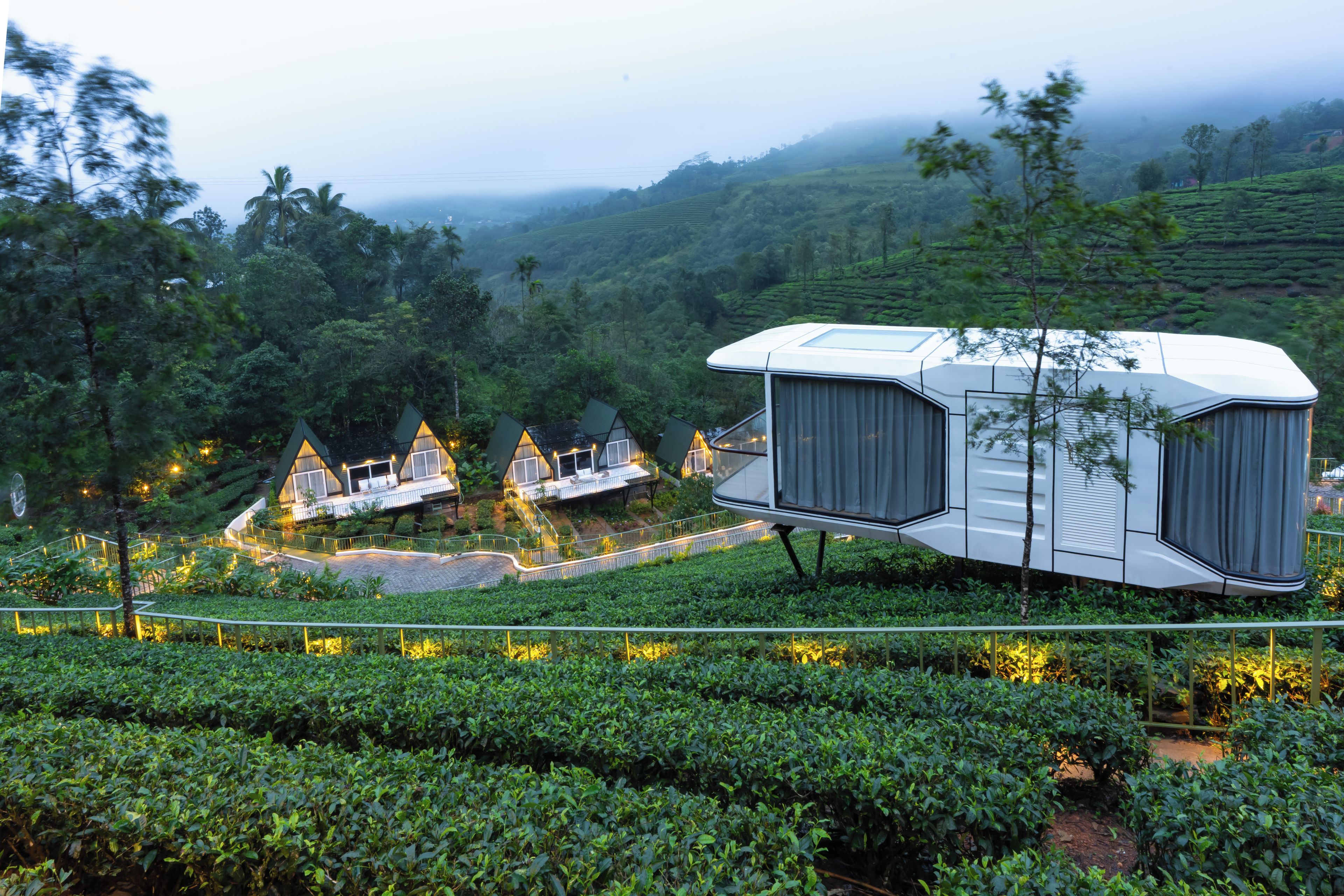 Una estructura moderna, blanca y de varias habitaciones con techo plano, rodeada de exuberante vegetación y plantaciones de té.