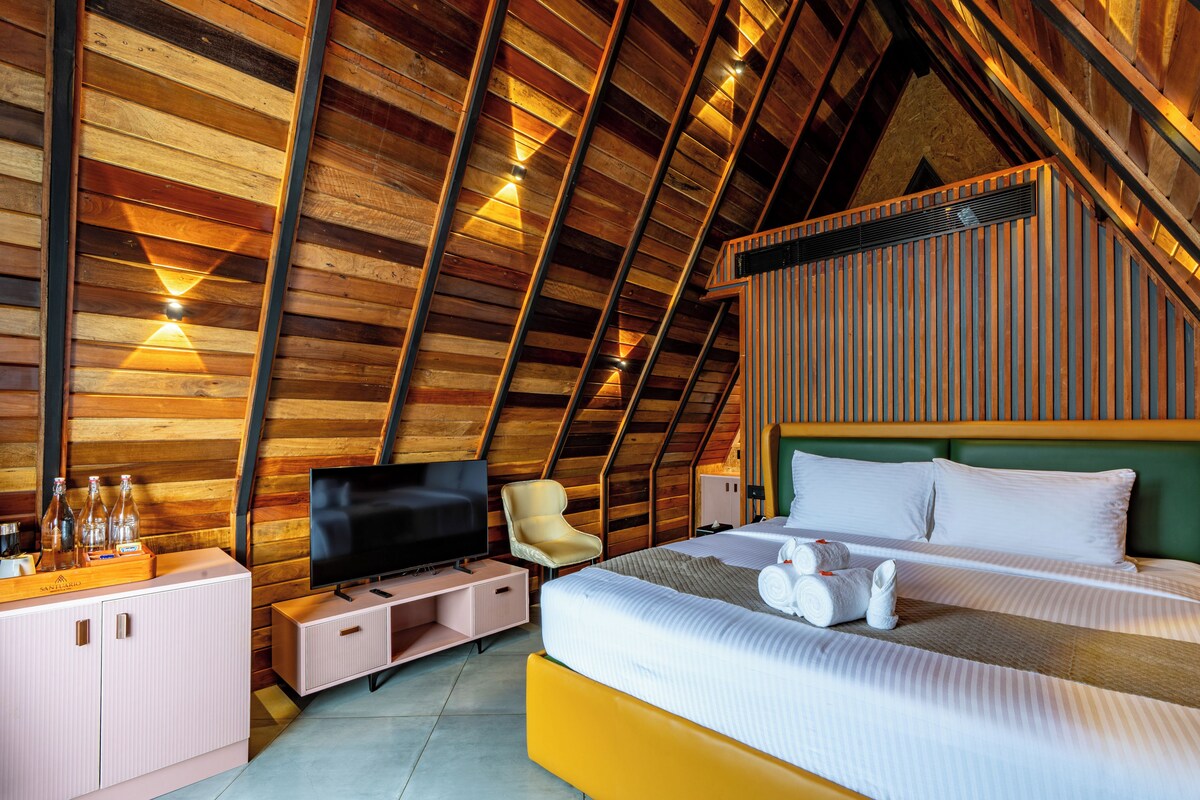 A room with a sloped ceiling, wooden paneling, a bed with white linens, a television, a chair, and a small table with bottles.