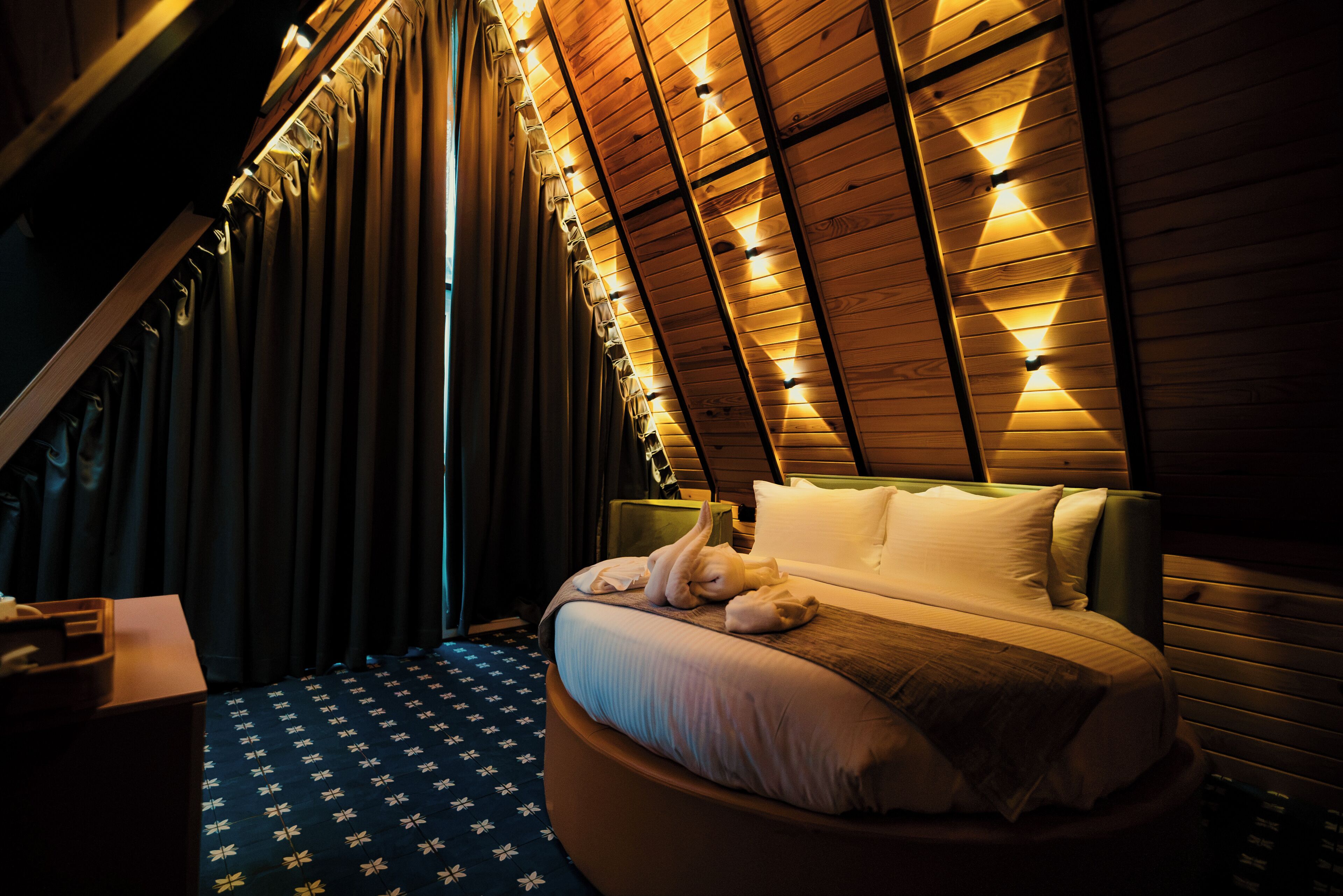 A bedroom with a sloped ceiling, a bed with pillows, a bedside table, and a patterned floor.