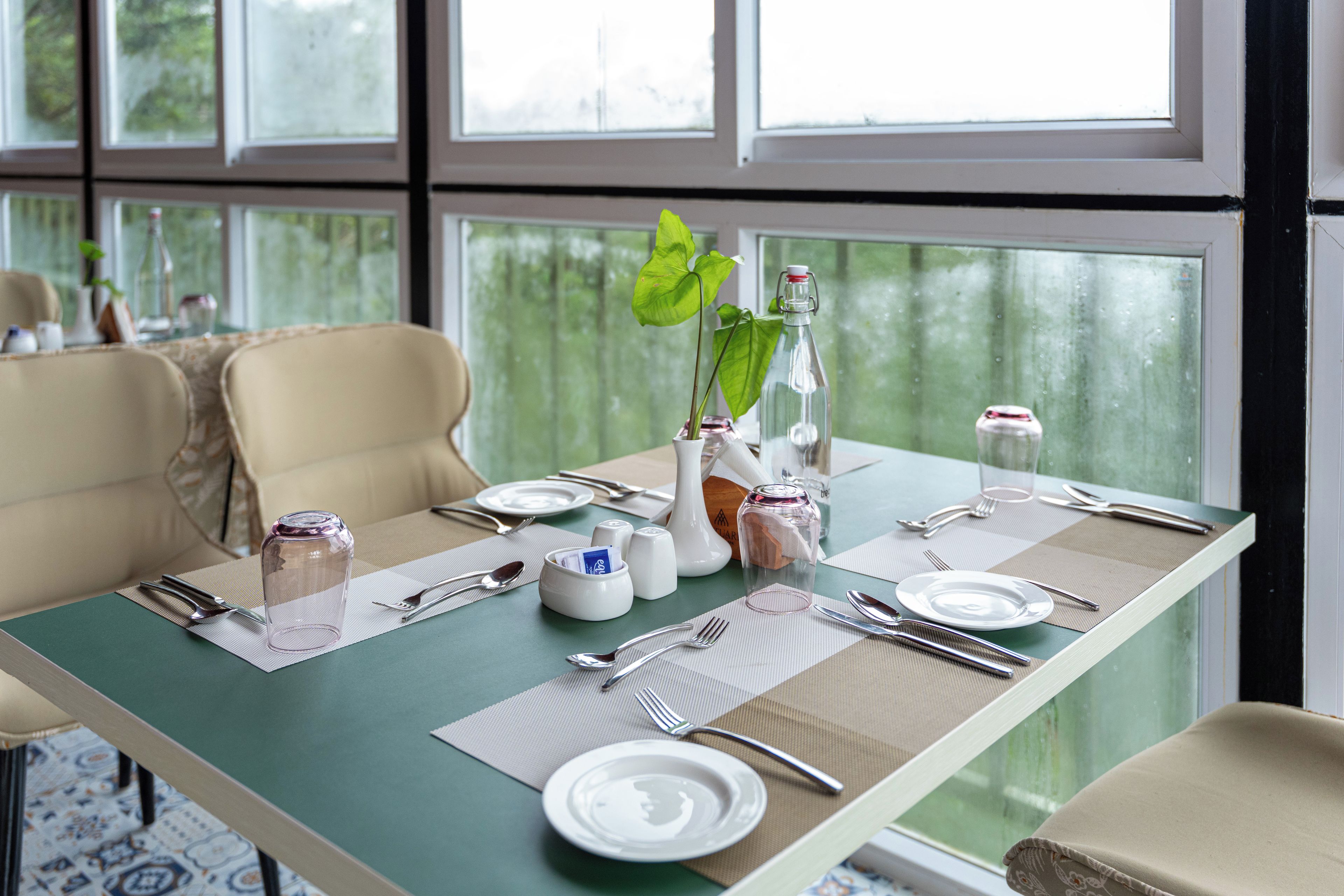 Mesa lista con platos, cubiertos y una botella, con vista a la vegetación por la ventana.