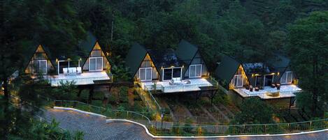 A row of triangular-roofed cabins with outdoor seating areas, surrounded by lush greenery.