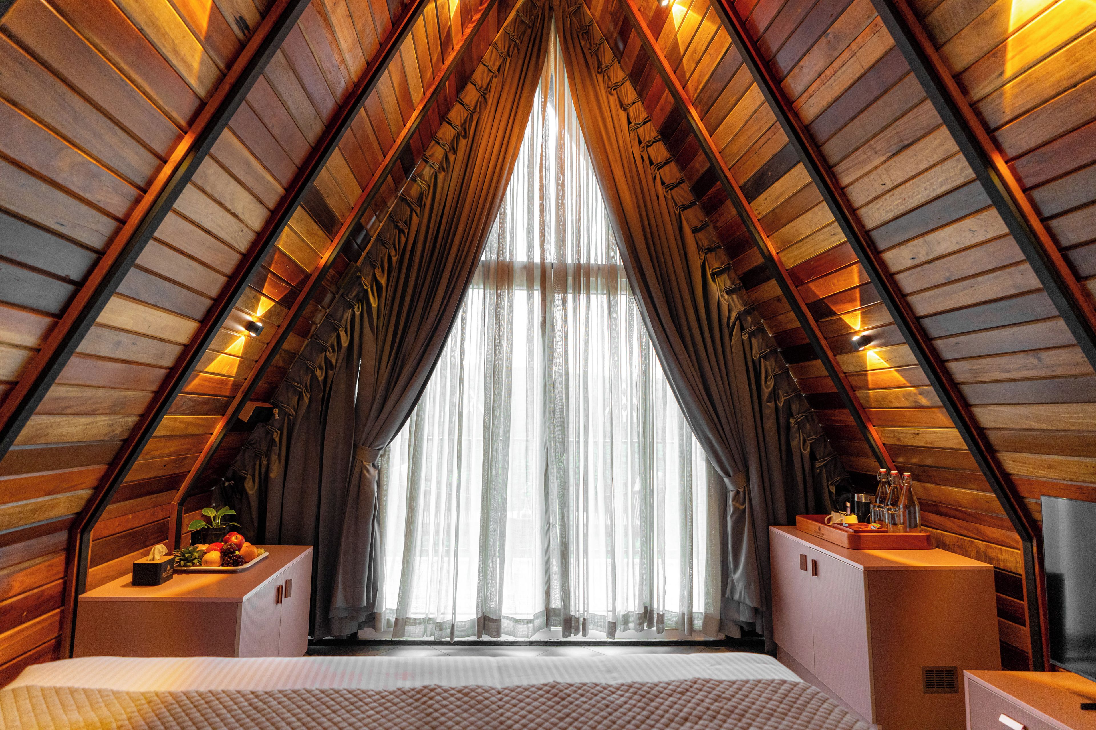 Une pièce aux hauts plafonds, dotée d’un plafond en bois incliné, d’une grande fenêtre ornée de rideaux légers et d’un lit avec un matelas blanc.