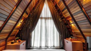 Une pièce aux hauts plafonds, dotée d’un plafond en bois incliné, d’une grande fenêtre ornée de rideaux légers et d’un lit avec un matelas blanc.