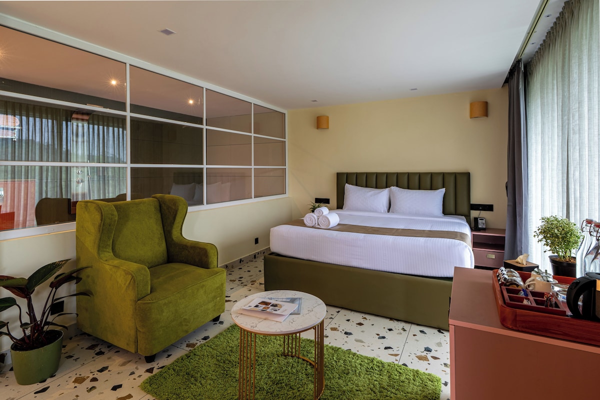 A hotel room with a bed, a green armchair, a small round table, and a potted plant.