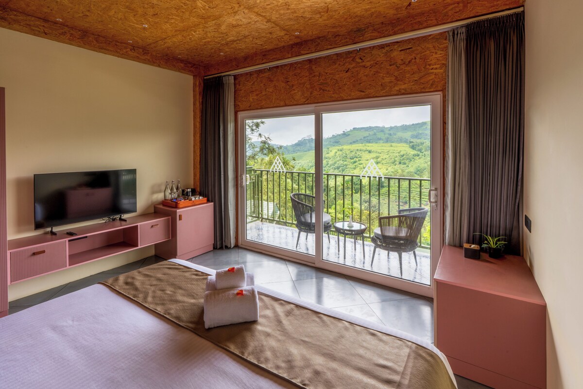 A room with a bed, a TV, a balcony with chairs, and a view of greenery.