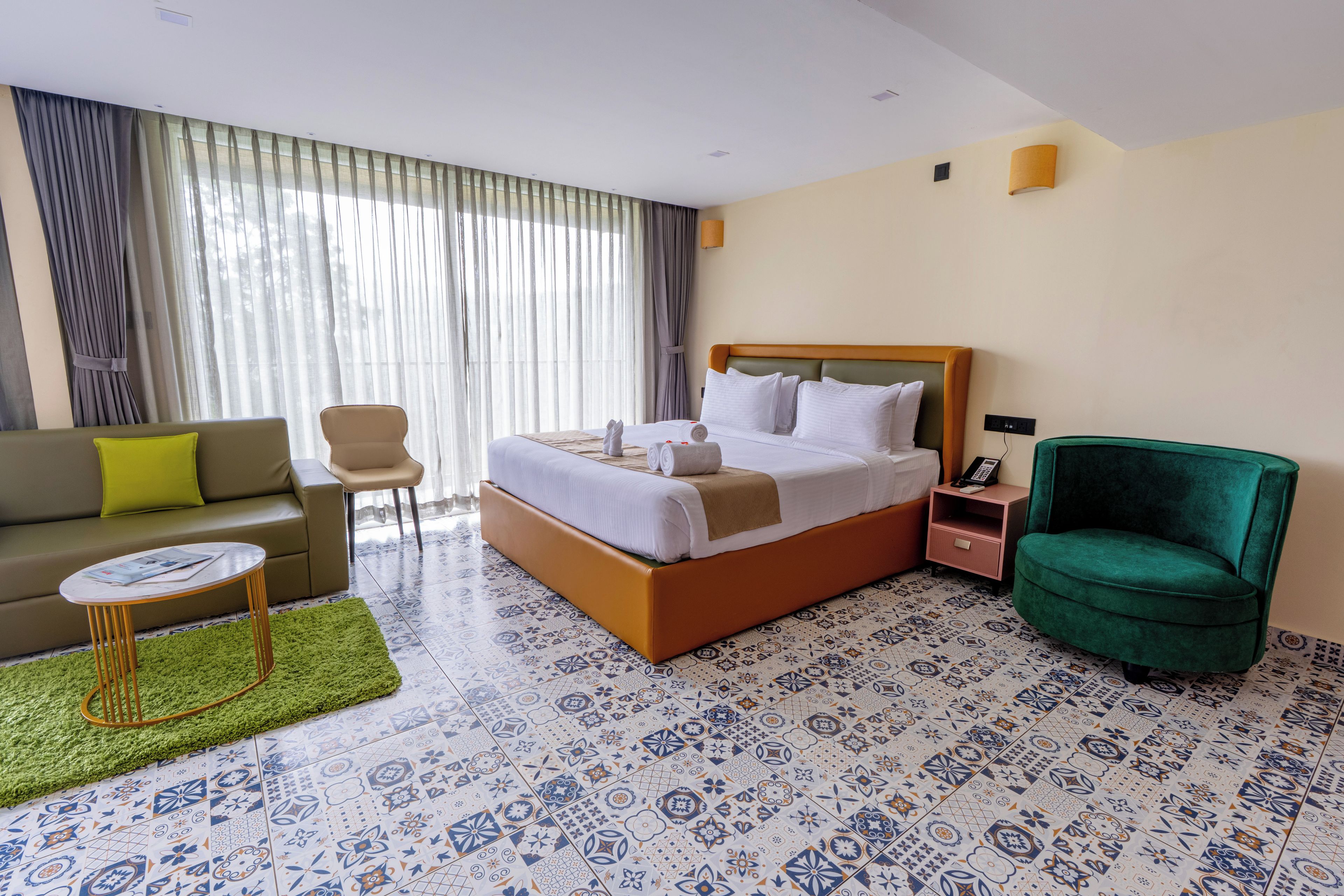 A hotel room with a bed, a sofa, a chair, a small table, and patterned floor tiles.