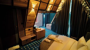 Una habitación con techo inclinado, una cama con ropa blanca, un aparador de madera y una mesita con una botella y vasos.