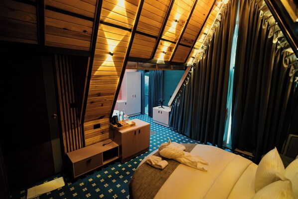 A room with a sloped ceiling, a bed with white linens, a wooden dresser, and a small table with a bottle and glasses.