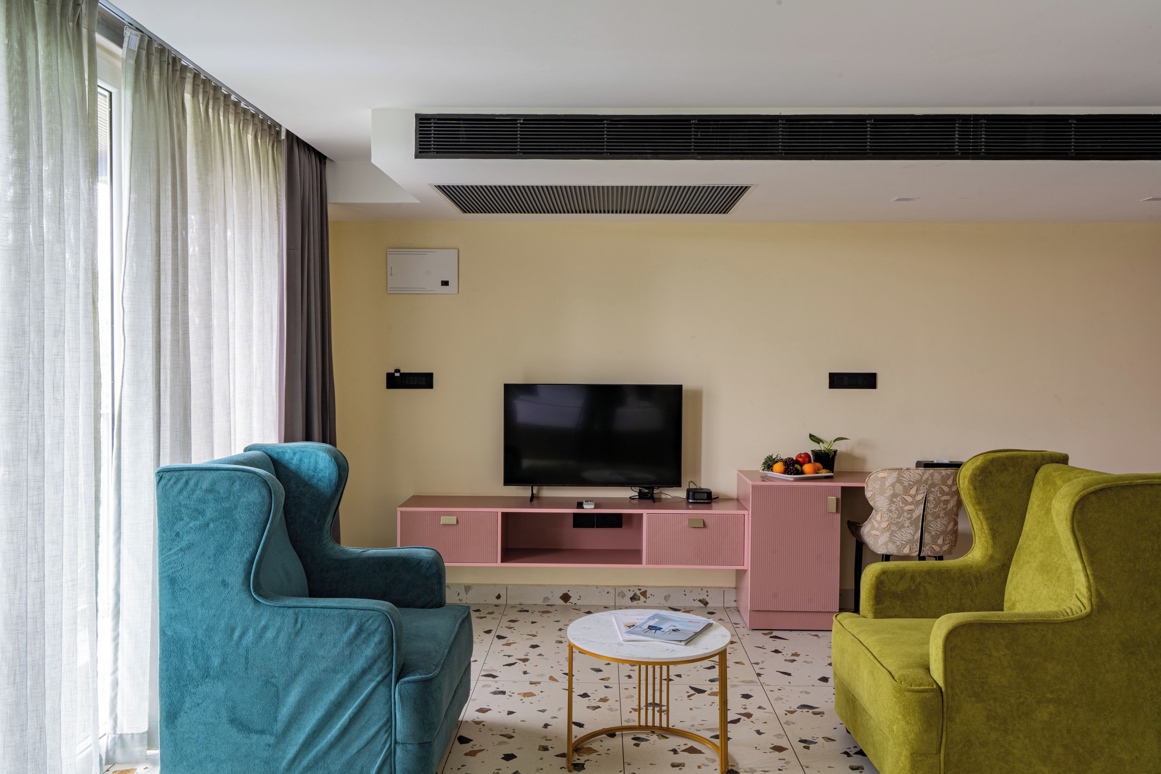 A living room with teal and green chairs, a pink entertainment unit, a flat-screen TV, and a small round table.