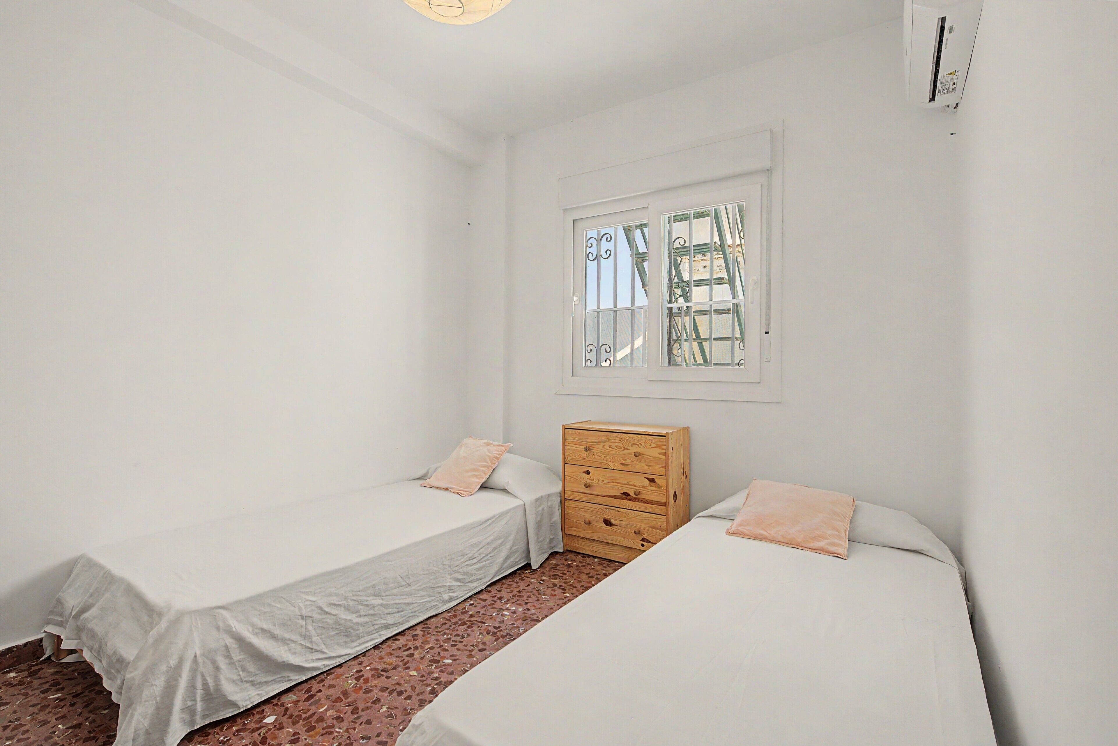 Una habitación pequeña y limpia con dos camas individuales, un aparador de madera y una ventana con barrotes.