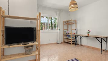 Ein Zimmer mit Fernseher auf einem Holzboden, einem Tisch, einer Hängeleuchte und einem Fenster mit Blick auf Grün.
