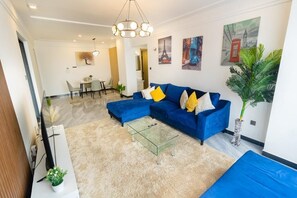 Ein modernes Wohnzimmer mit einem blauen Ecksofa, einem Glastisch und einem Essbereich im Hintergrund.