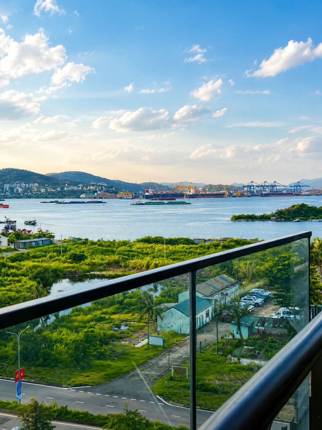 A view from a balcony overlooking a harbor with ships, greenery, and a distant cityscape.