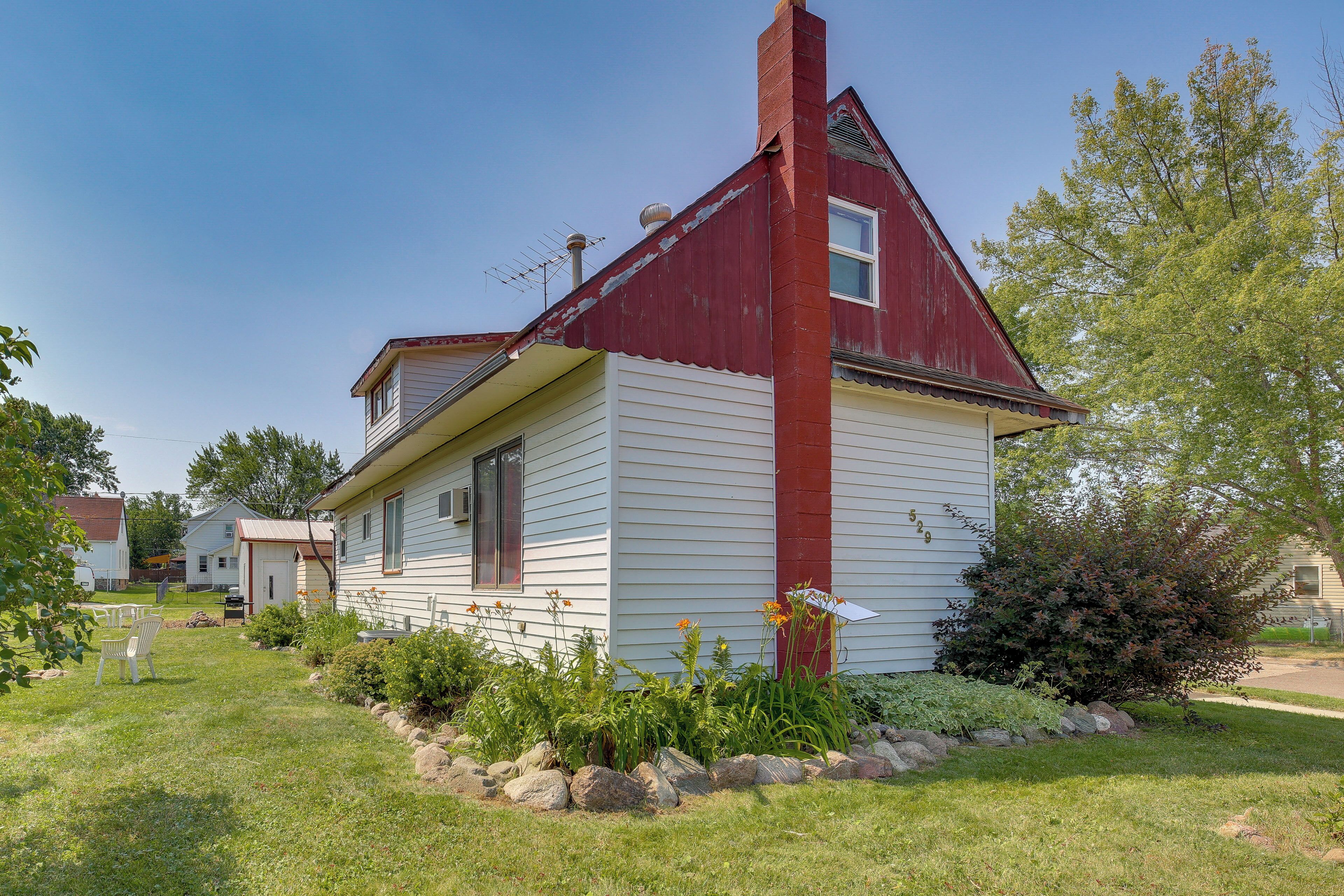 A single-story house with a red chimney, white siding, and a small front yard with rocks and plants.