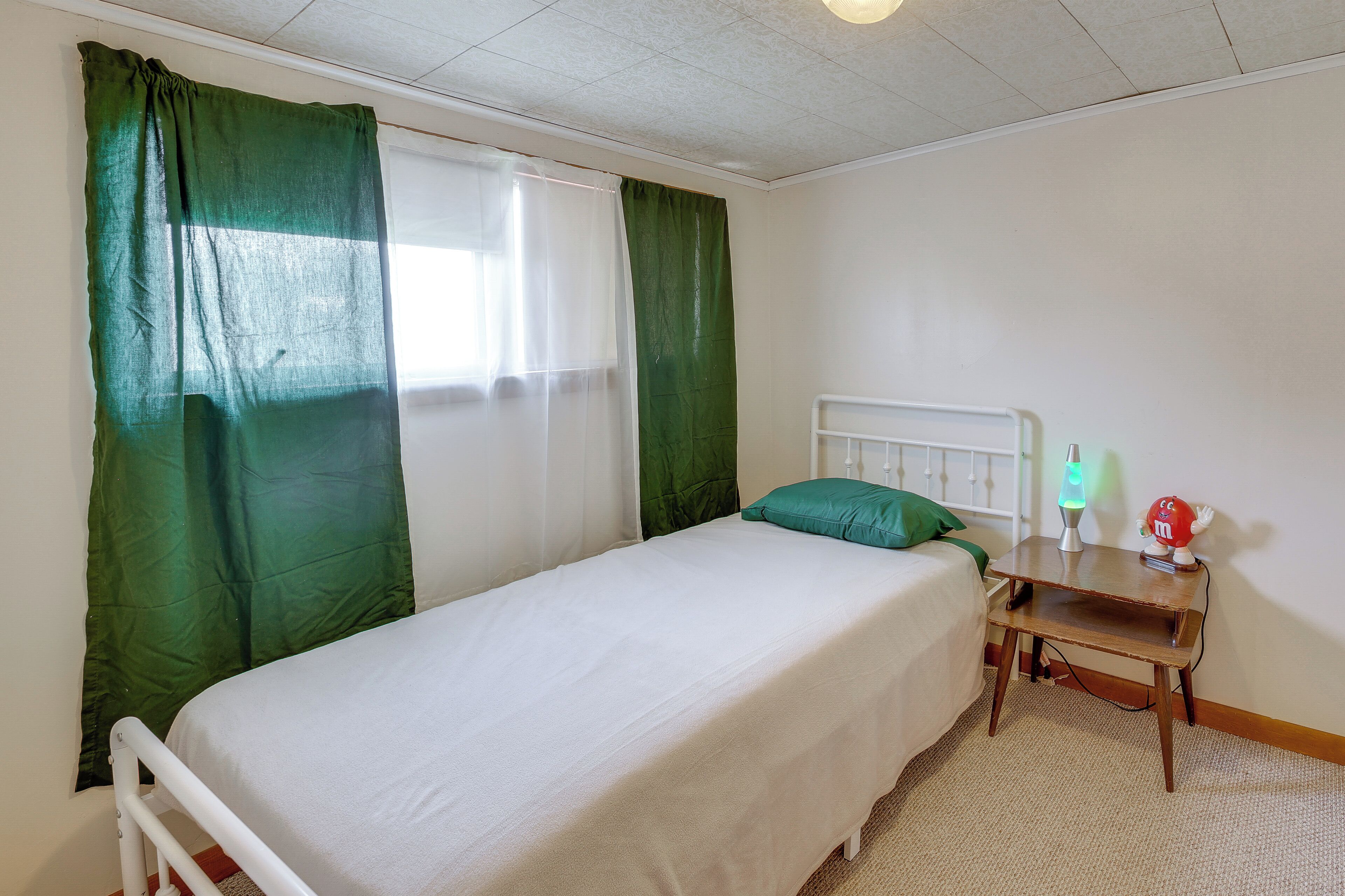 A single bed with a white mattress and green curtains, a bedside table with a lava lamp, and a window with sheer white curtains.