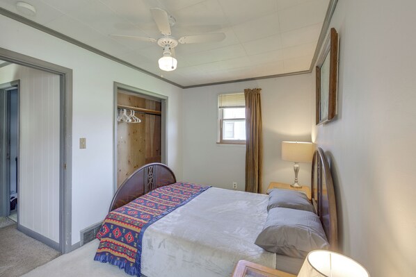 Un dormitorio con cama, ventilador de techo, ventana con cortinas y mesita de noche con lámpara.