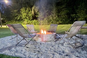 A fire pit surrounded by four chairs in a gravel area with a grassy field and trees in the background.