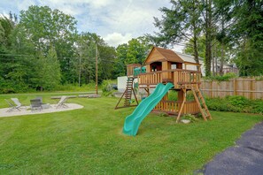 A wooden play structure with a slide, surrounded by a grassy yard and a wooden fence.
