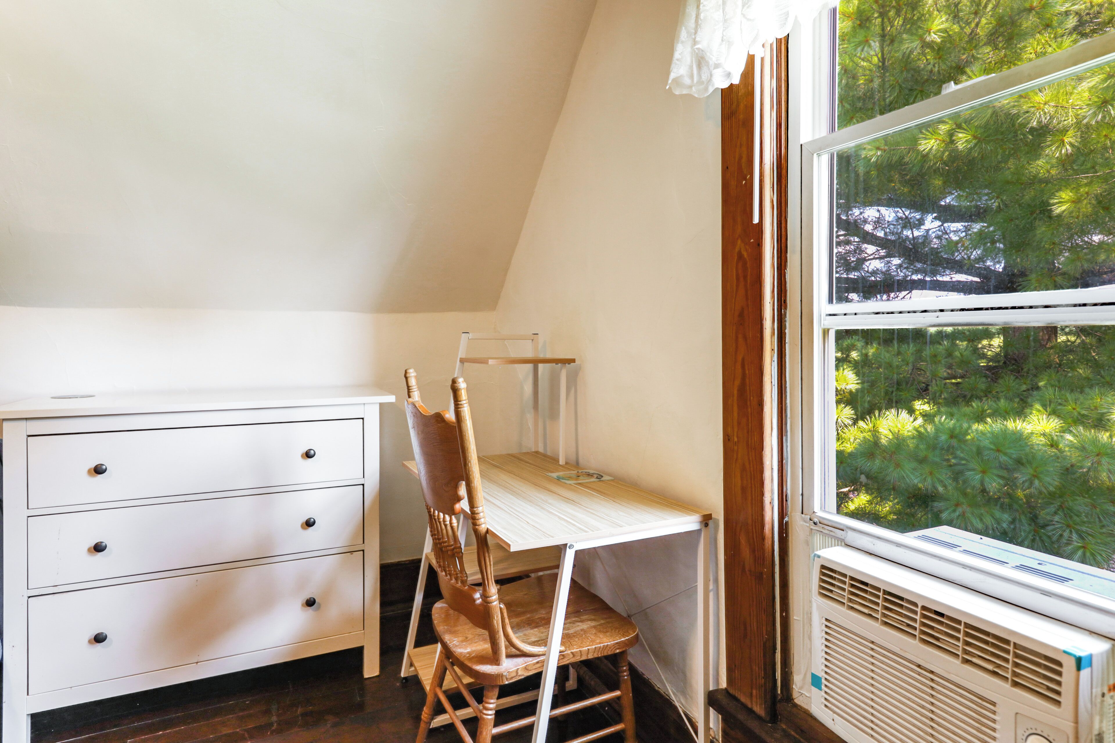 Une pièce avec une commode blanche, une chaise en bois, une petite table et une fenêtre donnant sur des arbres.