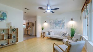 Un salon spacieux avec un canapé blanc, une chaise en bois, une table basse et un ventilateur de plafond. On y trouve une bibliothèque remplie de divers objets et un tableau accroché au mur.