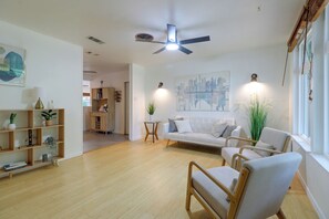 Ein geräumiges Wohnzimmer mit einem weißen Sofa, einem Holzstuhl, einem Couchtisch und einem Deckenventilator. Ein Bücherregal mit verschiedenen Gegenständen und ein Gemälde an der Wand sind ebenfalls vorhanden.