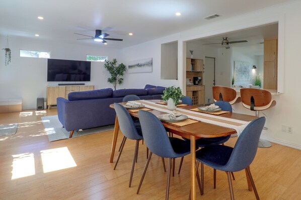 Een moderne woonkamer met een hoekbank, een eettafel met stoelen, een flatscreen televisie, plafondventilatoren en een groot raam.
