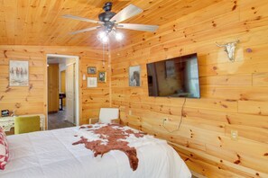 Ein Zimmer mit Holzwänden, einem Bett mit weißer und brauner Decke, einem Deckenventilator und einem an der Wand befestigten Fernseher.