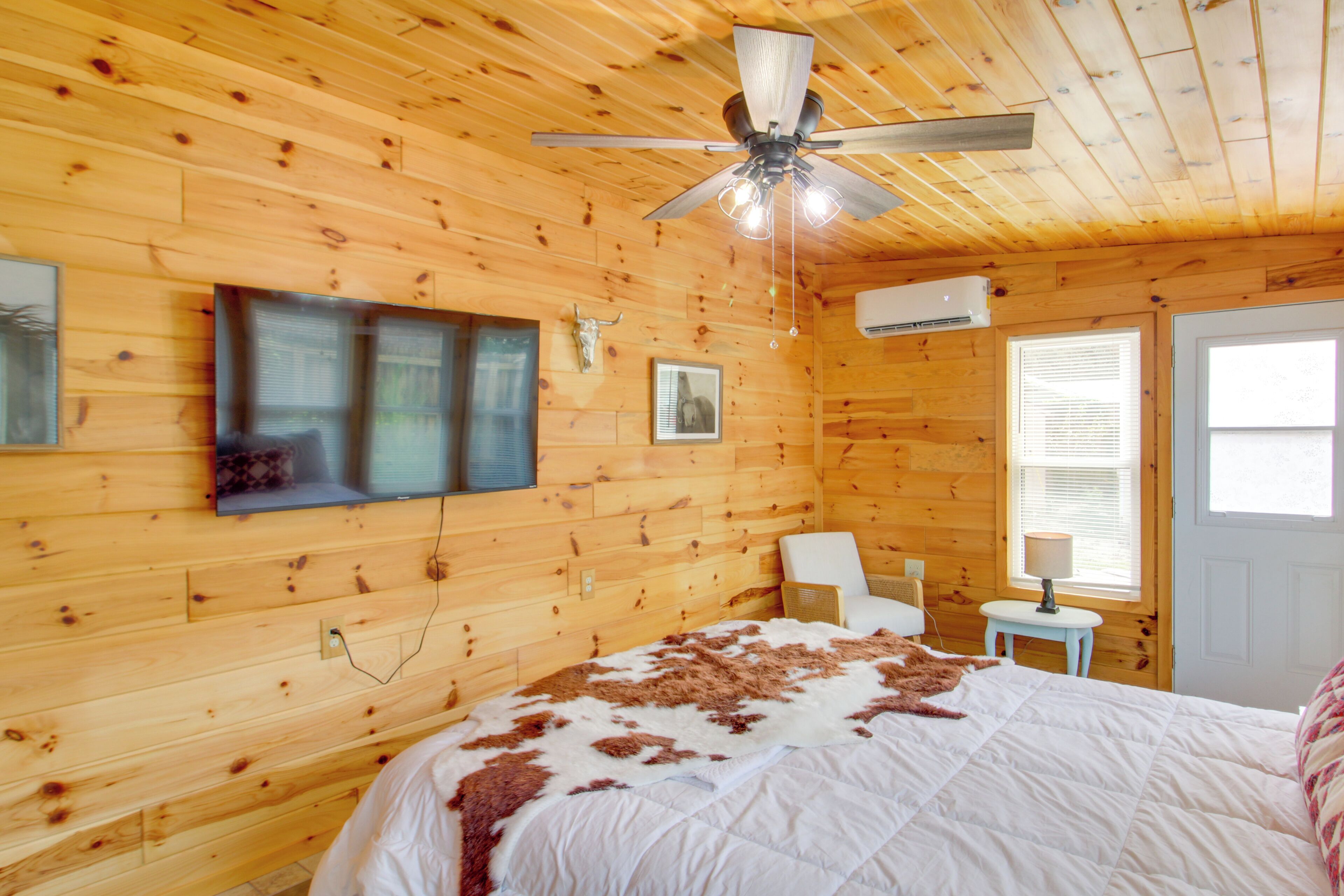 A wooden-paneled room with a bed, a ceiling fan, a mounted TV, and a window with blinds.
