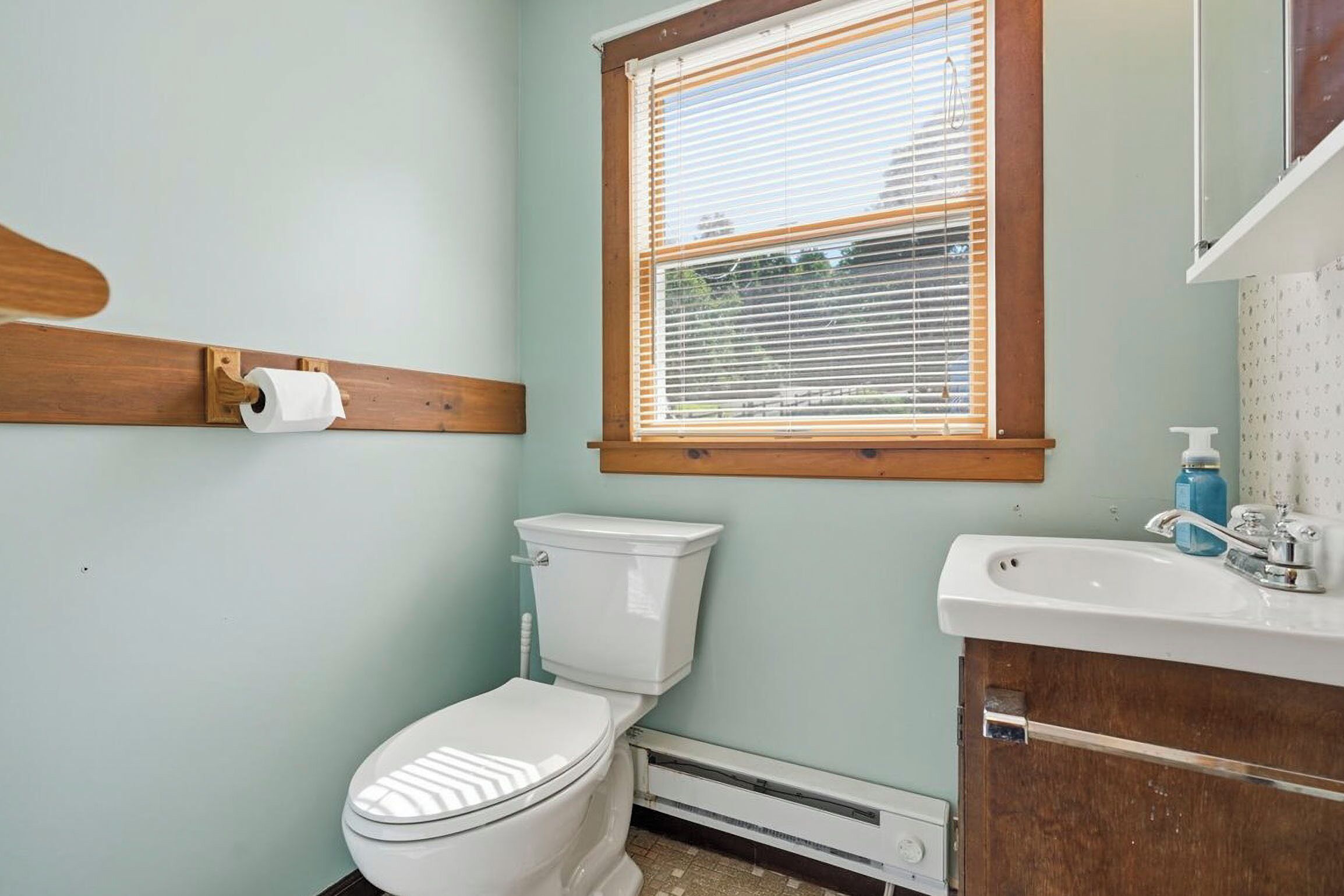 A bathroom with a toilet, a window with blinds, a roll of toilet paper, a sink, and a shower curtain.