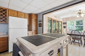 A kitchen with wooden cabinets, a white refrigerator, a dining table with chairs, and a view of the outdoors through large windows. - Lakefront Paradise on Lake Wallenpaupack w/ Dock (Hawley)