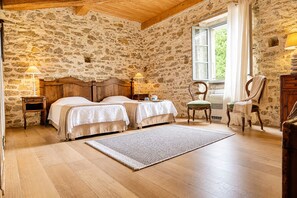 A bedroom with two beds, stone walls, wooden floors, a window with curtains, and a chair.