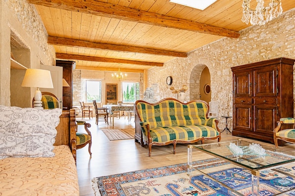 A spacious living room with a wooden ceiling, stone walls, a patterned sofa, a glass coffee table, and a dining area with chairs and a table.