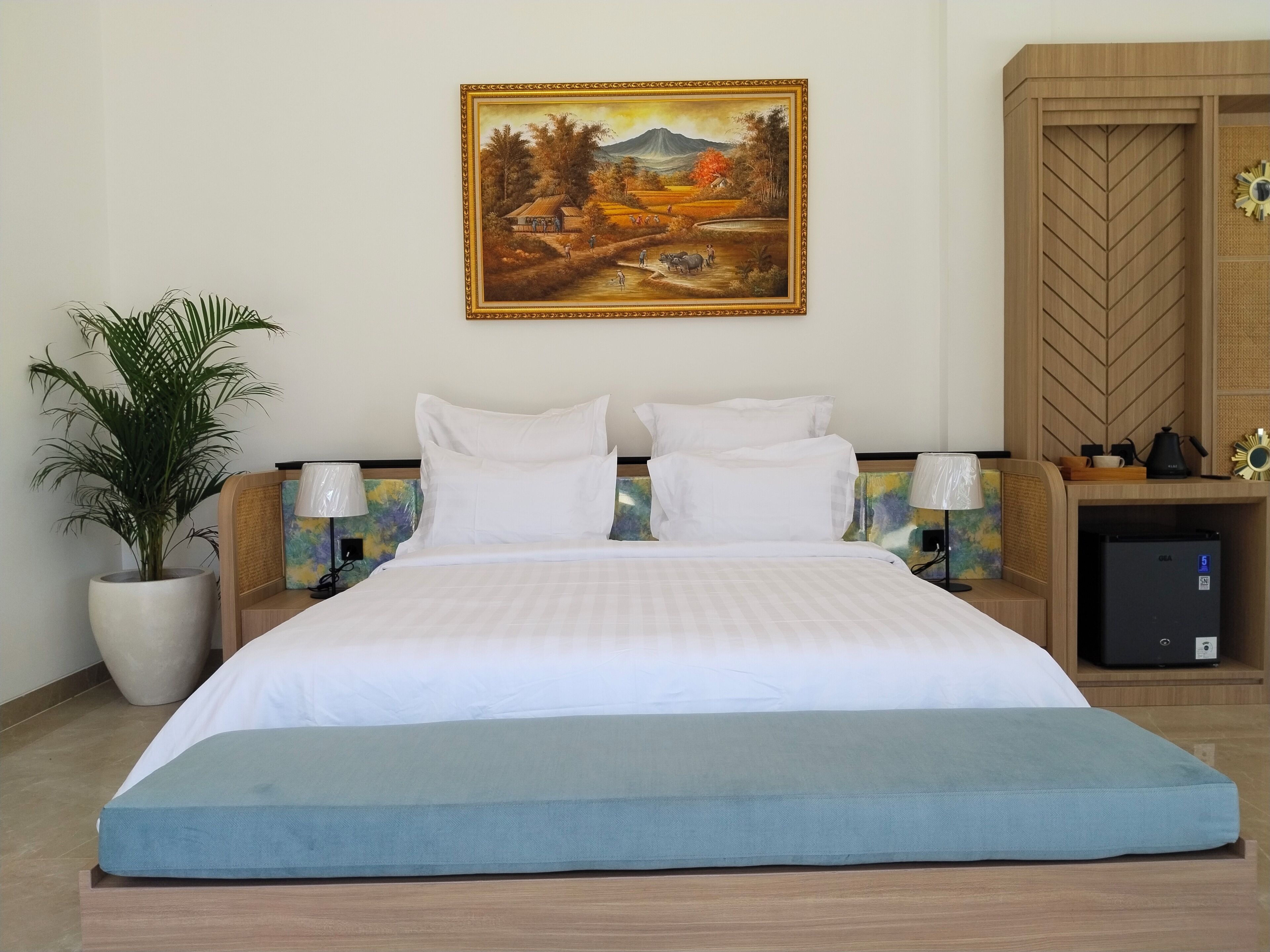 A hotel room with a bed, a nightstand, a lamp, a potted plant, and a painting on the wall.