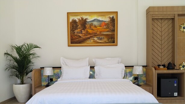 Habitación de hotel con cama, mesita de noche, lámpara, planta en maceta y un cuadro en la pared.