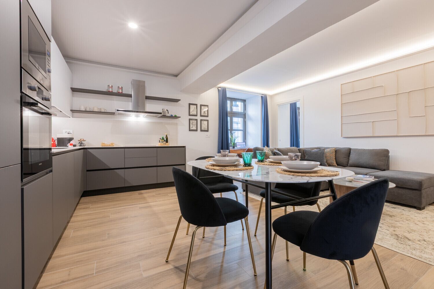 Et moderne køkken med indbyggede apparater, et spiseområde med et rundt bord og stole, samt en opholdsstue med sofa og et stort abstrakt kunstværk på væggen.