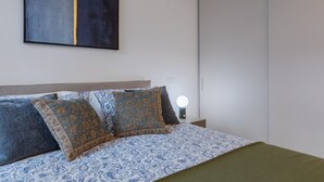 Ein Bett mit gemusterten Betttextilien, ein Nachttisch mit Lampe und ein an der Wand hängendes abstraktes Gemälde.