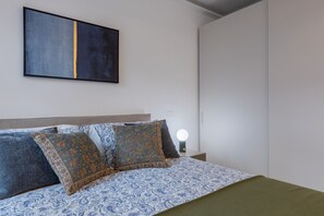 Ein Bett mit gemusterten Betttextilien, ein Nachttisch mit Lampe und ein an der Wand hängendes abstraktes Gemälde.