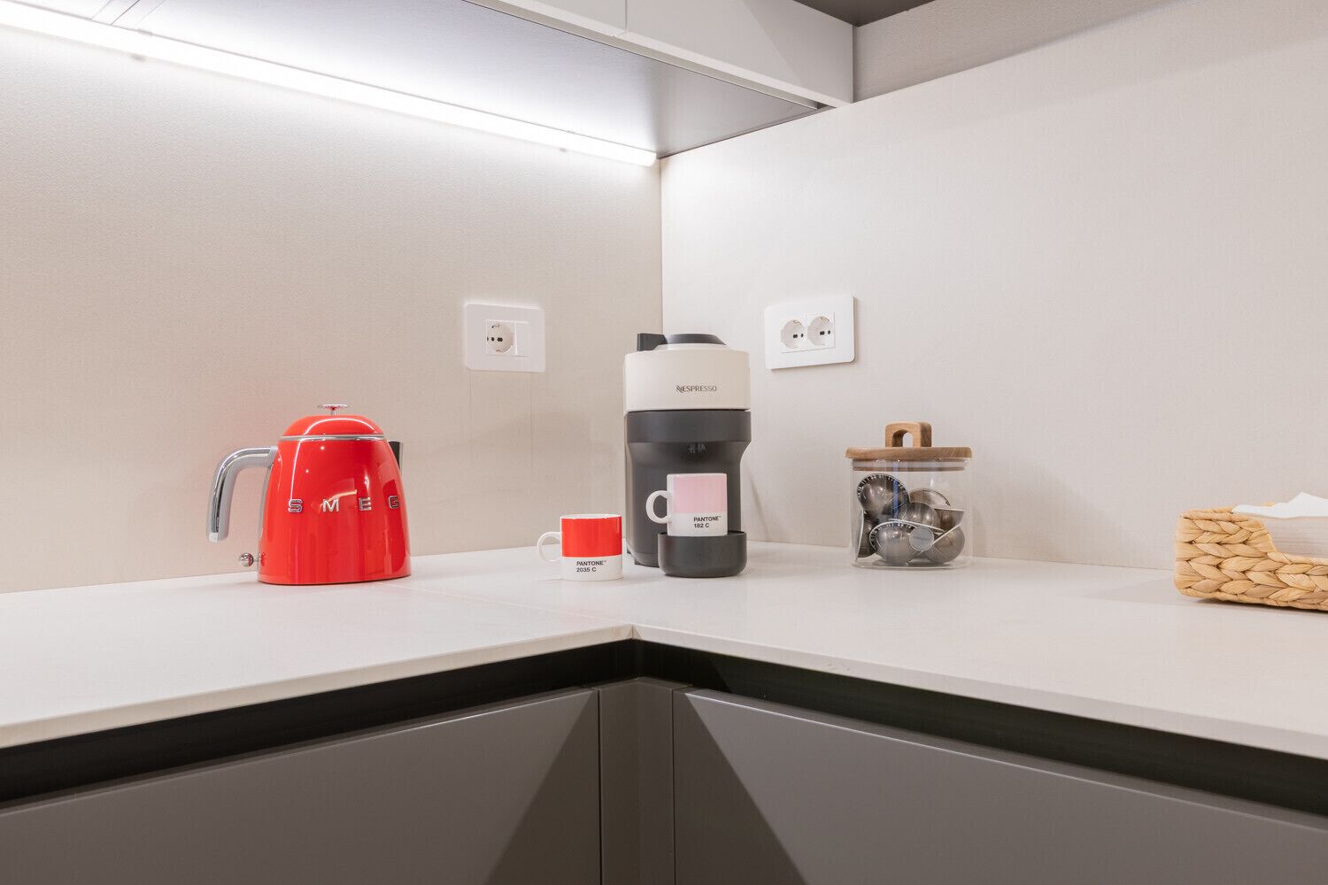 Una encimera de cocina con una cafetera roja, una máquina Nespresso, una taza y un tarro con utensilios.