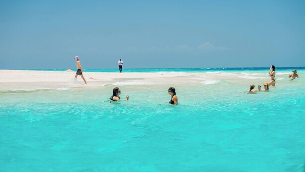 Une scène de plage avec une eau turquoise et limpide, des gens qui nagent et qui se promènent sur le sable.