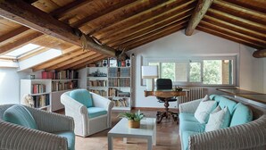 Um amplo loft com pé direito alto, vigas de madeira aparentes, um grande claraboia, uma área de estar aconchegante com móveis de vime e uma estante repleta de livros.