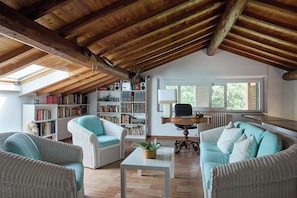 Ein geräumiges Loft mit hohen Decken, freiliegenden Holzbalken, einem großen Oberlicht, einer gemütlichen Sitzecke mit Korbmöbeln und einem Bücherregal voller Bücher.